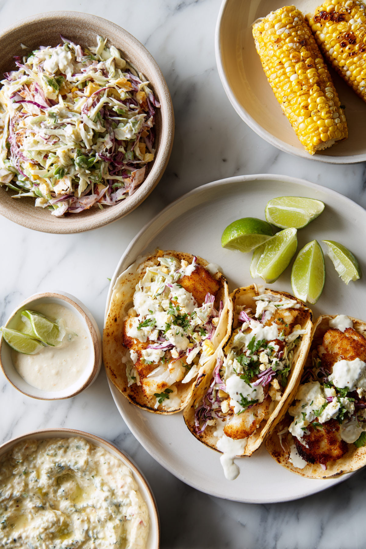 fish tacos served with additional slaw and toppings on the side and a side of Mexican street corn.