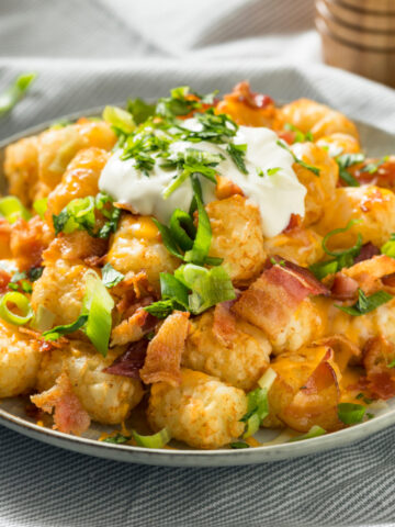 A plate of tater tots topped with melted cheese, bacon, sliced green onions, and sour cream.