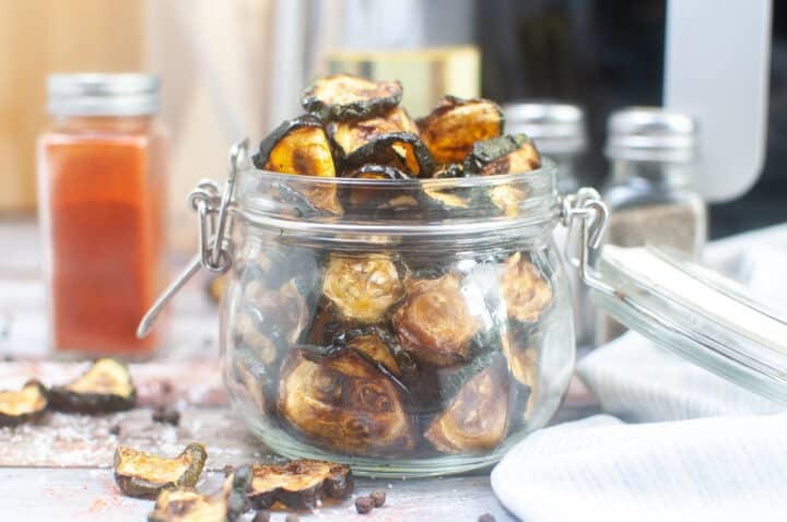Side view of air fried zucchini chips in a glass jar with the seasoning ingredients in the background