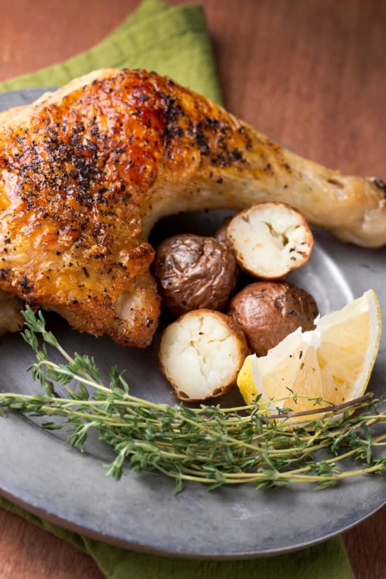 crispy roasted chicken leg quarter on a plate served with roasted red potatoes and garnished with lemon wedge and rosemary