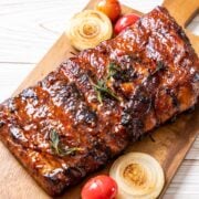 St Louis style pork ribs baked, glazed and broiled. Ribs are laying on a wooden board with roasted sliced onions and tomatoes alongside