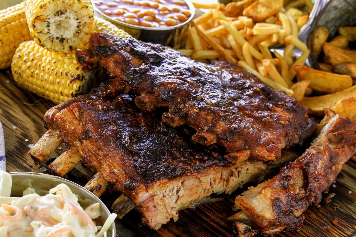 slow cooked bbq beef back ribs displayed on a wooden board with coleslaw, roasted corn on the cob, French fries, potato wedges and baked beans. a full course beef bbq rib dinner is shown.