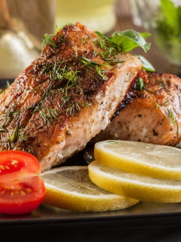 2 Broiled salmon fillets served on a dark plate with lemon slices and halved small tomatoes as garnish. Side view