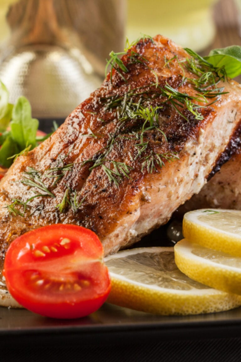 Broiled salmon fillets served on a dark plate with lemon slices and halved small tomatoes as garnish. Side view