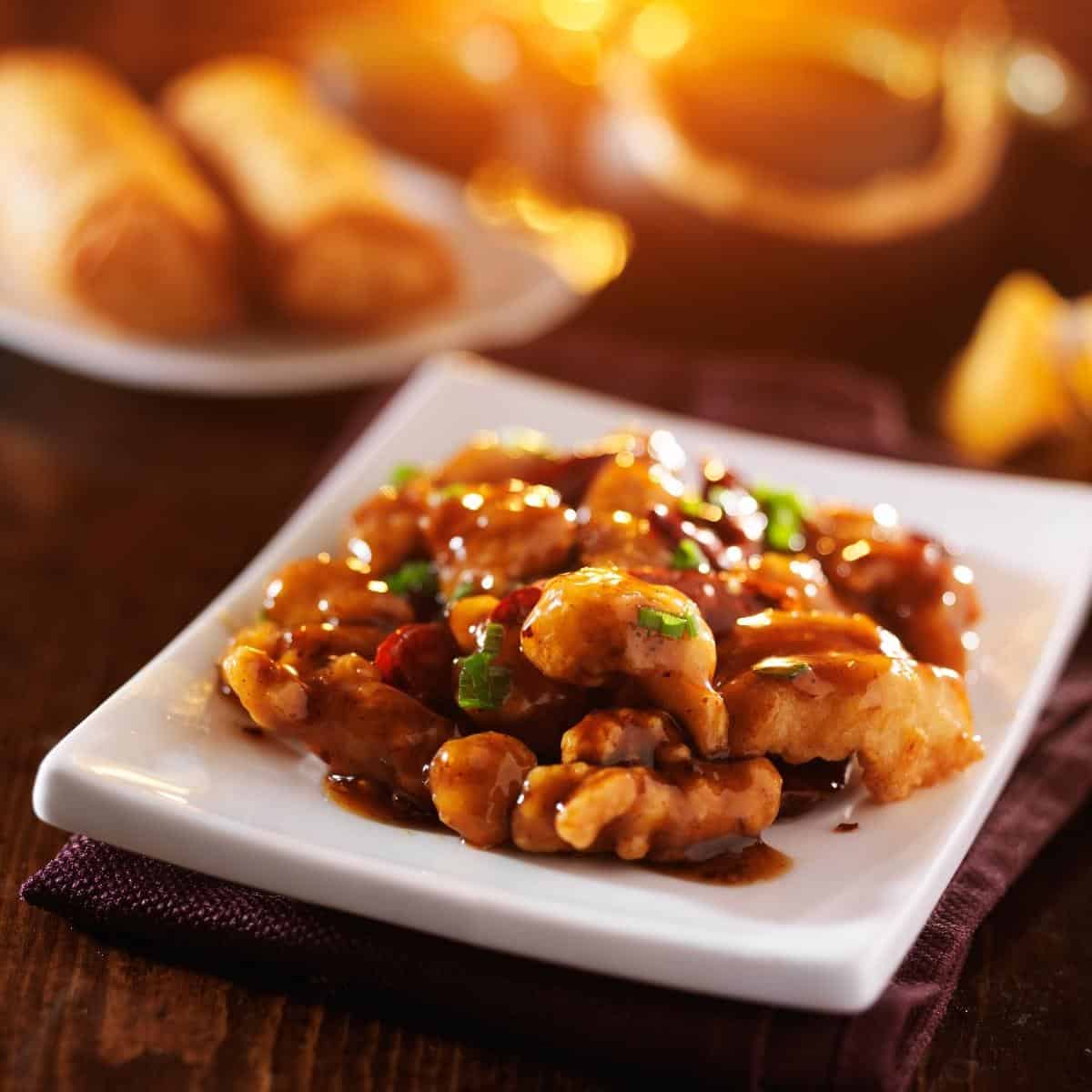 general tso's chicken served on a square white plate with egg rolls in the background