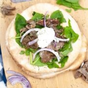 gyro sandwich top down view on a wood carving board. pita bread is topped with lettuce, shaved and seasoned beef, sliced red onion and tzatziki sauce.