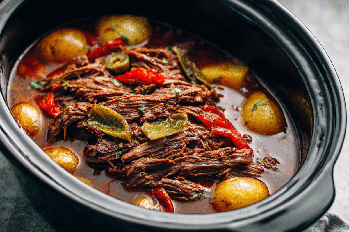 Shredded chuck roast with pepperoncini peppers and potatoes in a slow cooker insert on a light stone countertop