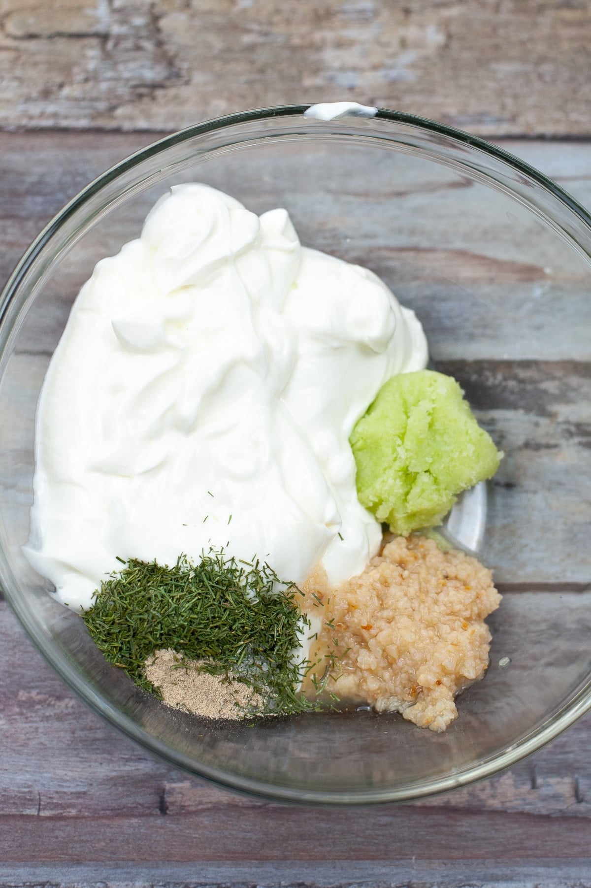 ingredients for tzatziki sauce in a clear bowl showing the sour cream, herbs and drained cucumber mash