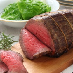 Carved rump roast on a wood carving board with a bowl of greens ready to be served.