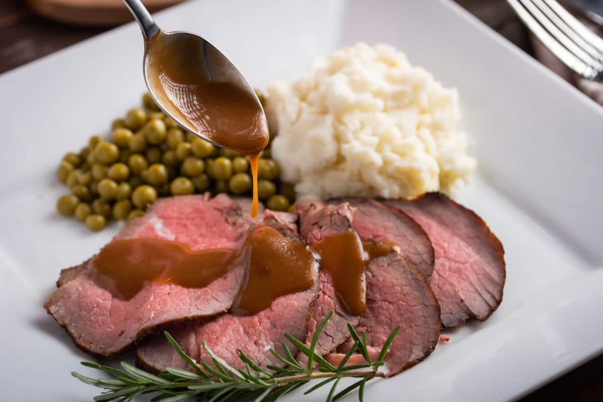 sirloin tip roast carved on a plate served with potatoes and peas. gravy drizzled over roast with a spoon.