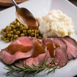 sirloin tip roast carved on a plate served with potatoes and peas. gravy drizzled over roast with a spoon.