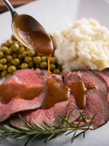 sirloin tip roast carved on a plate served with potatoes and peas. gravy drizzled over roast with a spoon.