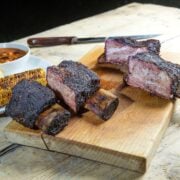 four smoked beef ribs on a cutting board with grilled corn and baked beans in the background