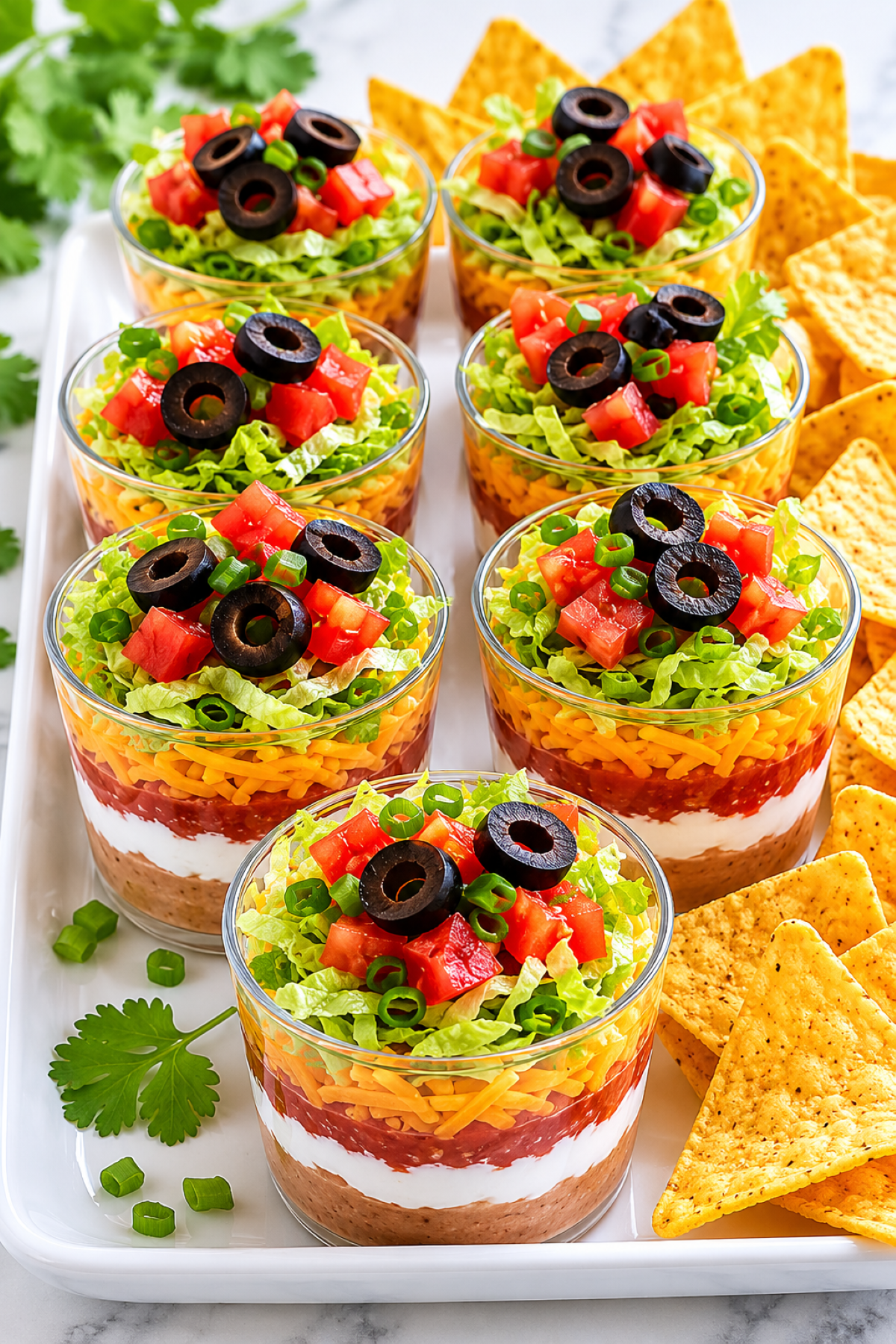 individual appetizer cups of 7 layer dip on a platter with chips served alongside